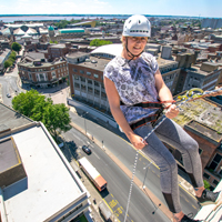 Photograph looking down at someone abseiling down a building with a view of the city in the background