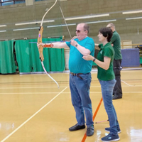 Photo of someone aiming a bow and arrow and being instructed by a spotter